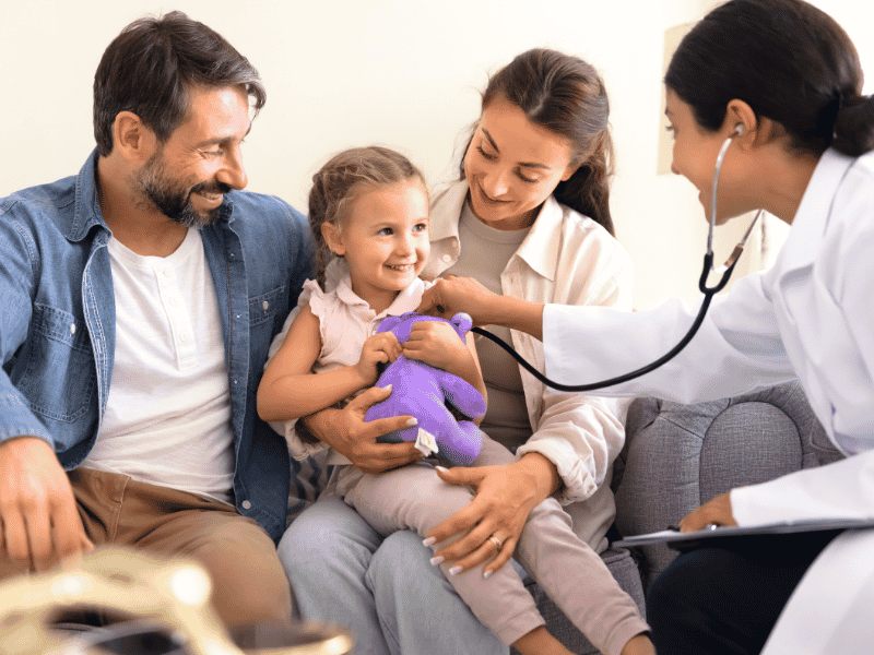 Female family doctor listening to a young girl's chest with a stethoscope while her parents sit beside her during a GP visit